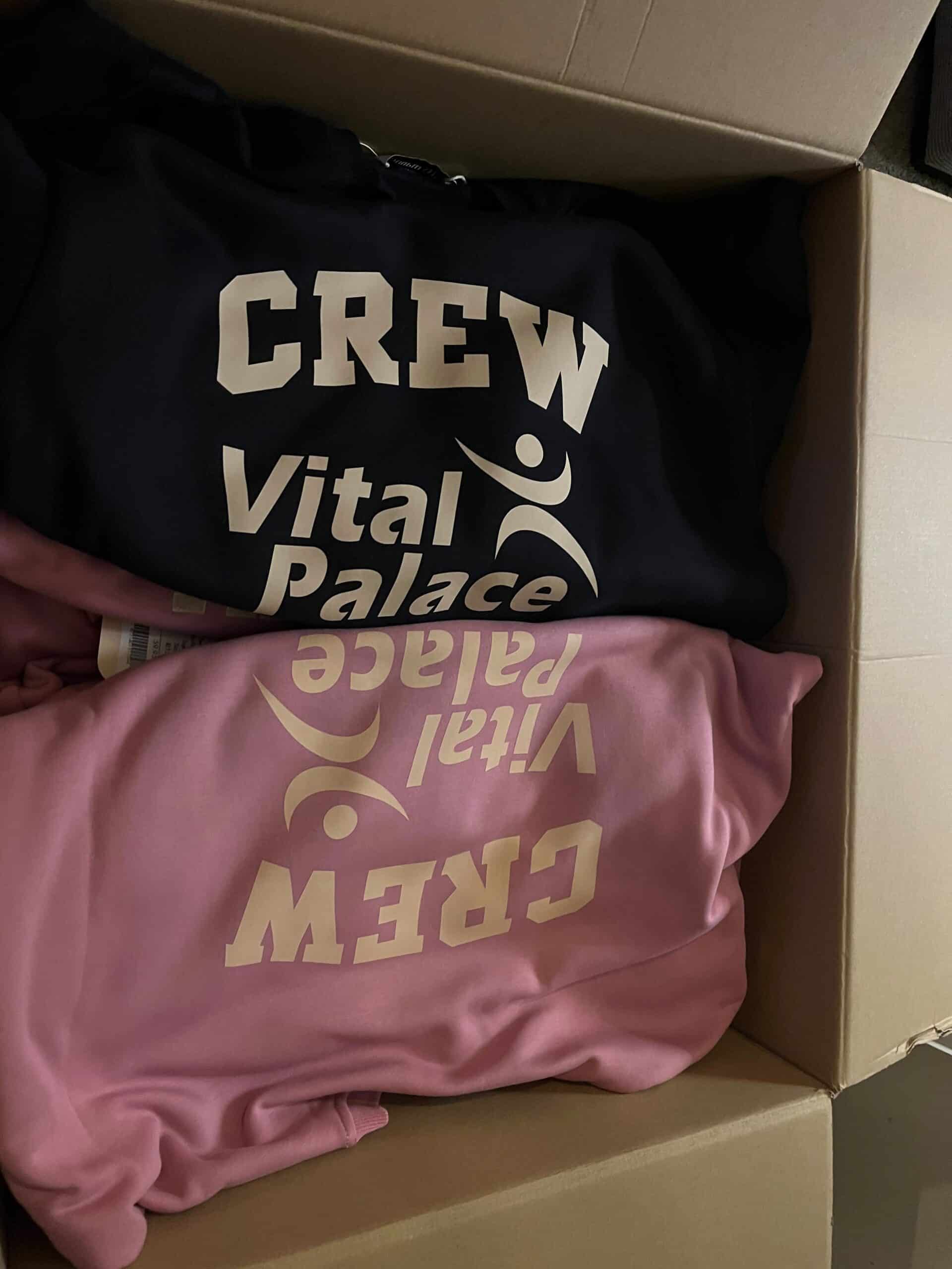 Roze en donkere truien met de CREW Vital Palace-bedrukking in een doos.